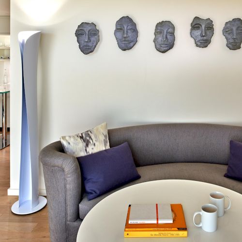 Five sculpted faces wall-art above a gray couch; a round coffee table with books and mugs; cozy living room with purple cushions.