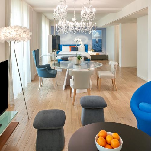 Bright, modern living room with a chandelier, round dining table, mix of blue and gray seating, and a bowl of oranges on a dark table.
