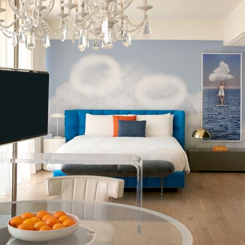 A bright ocean‑themed hotel room with a blue upholstered bed, white bedding, a glass table, flat-screen TV, chandelier, and a bowl of oranges.