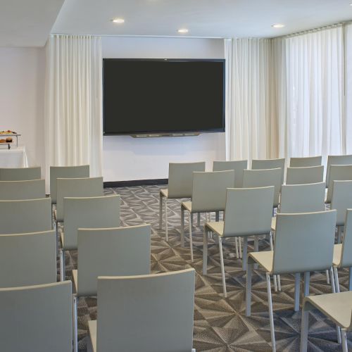 A modern conference room with white chairs facing a large wall-mounted screen, soft curtains, and a small refreshment table to the left.