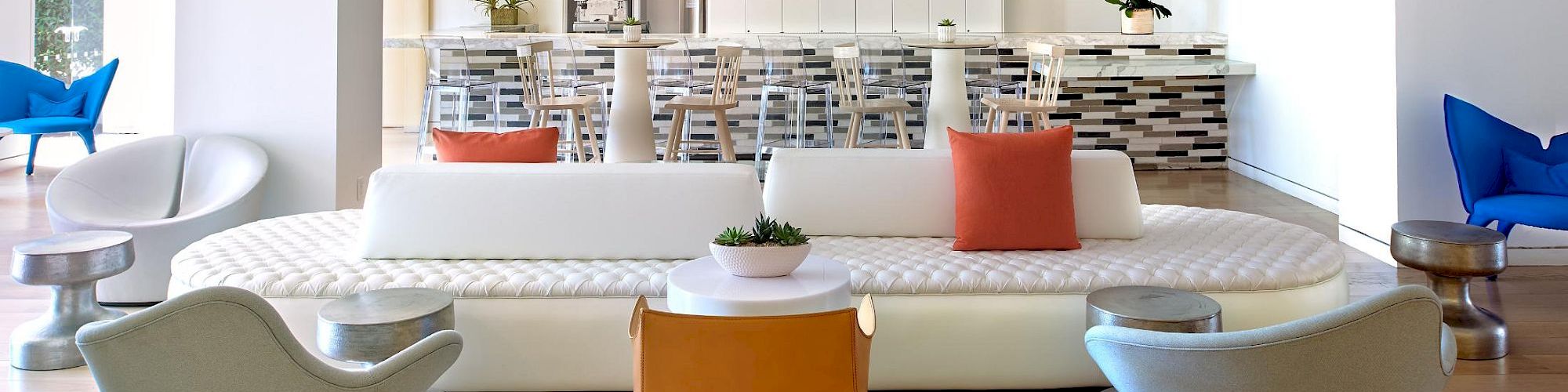 A bright, open living area with a white sofa, orange throw pillow, modern chairs, and a sleek kitchen island in the background, 140 characters ending with a period.
