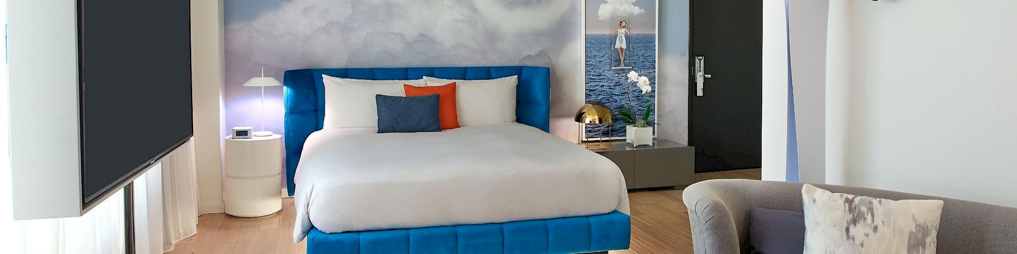 A modern hotel room with a blue-themed bed, sky mural, circular lights, a sofa and ottoman, round table with a notebook, and wall masks near the doorway.