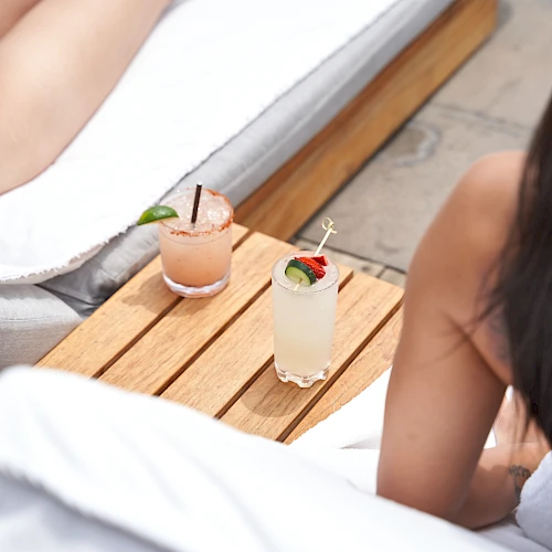 Two people relaxing by a pool with drinks on a wooden table; towels and lounge chairs, summery vibe.